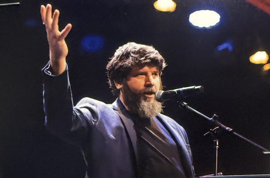 Tom at a microphone on stage with one arm raised