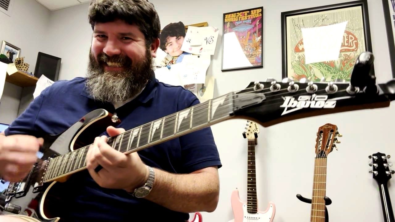 Tom smiling and playing an electric guitar in his teaching room, guitars hanging on the wall behind him